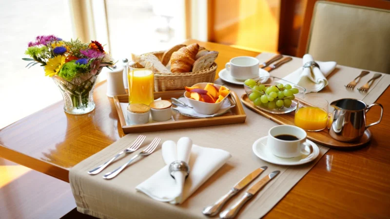 Fotografia realista de uma mesa posta para café da manhã, com trilho bege, xícaras de porcelana brancas, cesta de pães rústica, guardanapos de tecido com argolas, arranjo floral, talheres inox e bandeja de bambu, sob luz natural.