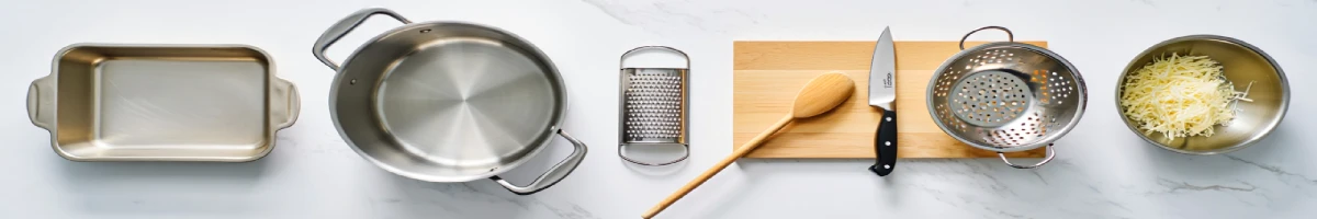 Utensílios de cozinha organizados em bancada: assadeira, panela, ralador, tábua de corte com faca e colher de madeira, peneira e tigela com queijo ralado.