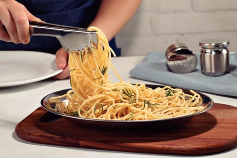 Pessoa utilizando pegador de massas em aço inox para servir espaguete com ervas em prato de metal sobre tábua de madeira.