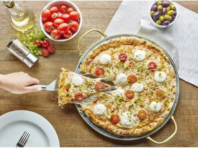 Pedra para pizza Electrolux com alças douradas sendo usada para servir pizza de mussarela de búfala com tomates-cereja sobre mesa rústica de madeira